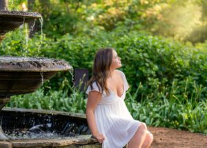 White dress portrait by garden fountain in warm sunset lighting with blooming greenery in background.