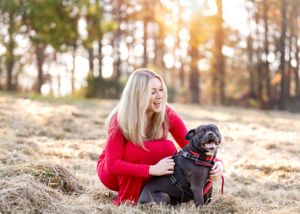 Person in red dress kneeling with black dog in sunny autumn field.