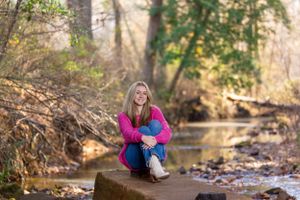 Person in bright pink sweater sitting on rock by stream in autumn forest.