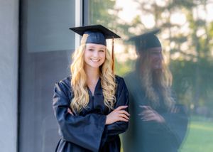 Graduate wearing navy blue academic regalia poses against reflective glass surface.