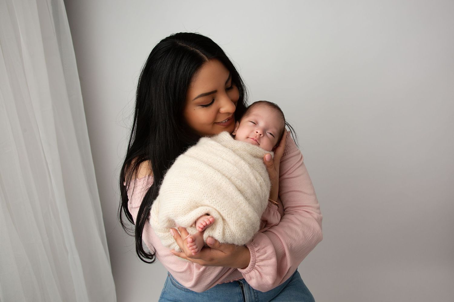 A gentle embrace between mother and newborn baby swaddled in white against a neutral background.