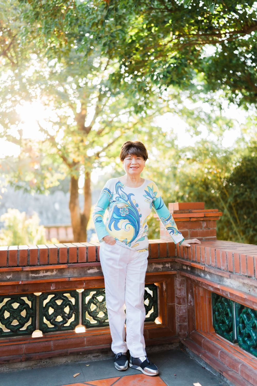 Person in white pants and patterned blue top stands on brick terrace under sunlit trees.