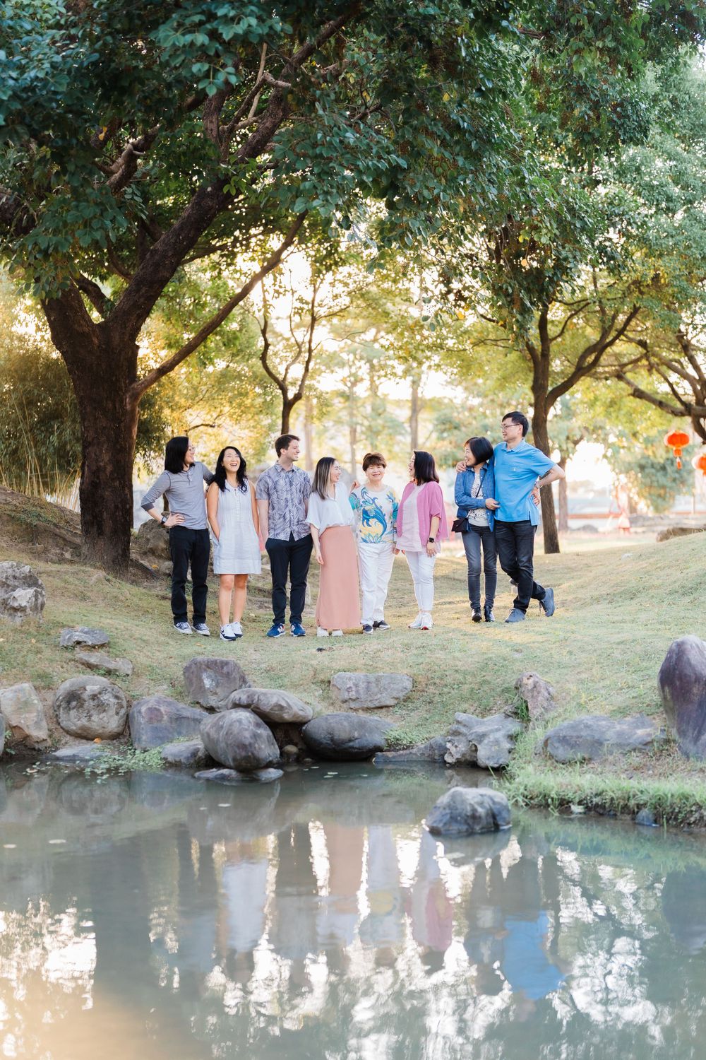 A group stands near a reflective pond under large trees during golden hour.