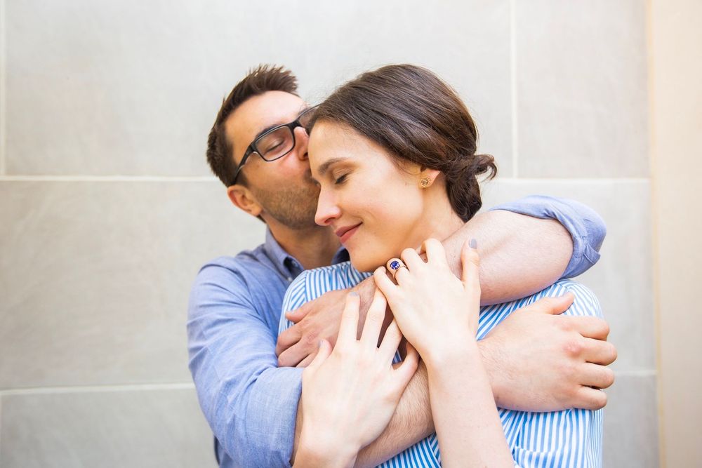 Affectionate man kissing woman on cheek while embracing her from behind against tiled wall.