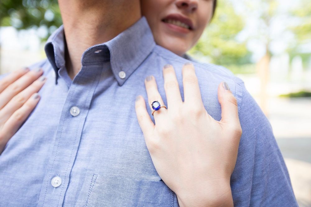 Close-up of woman's hand with blue sapphire engagement ring resting on man's light blue shirt.