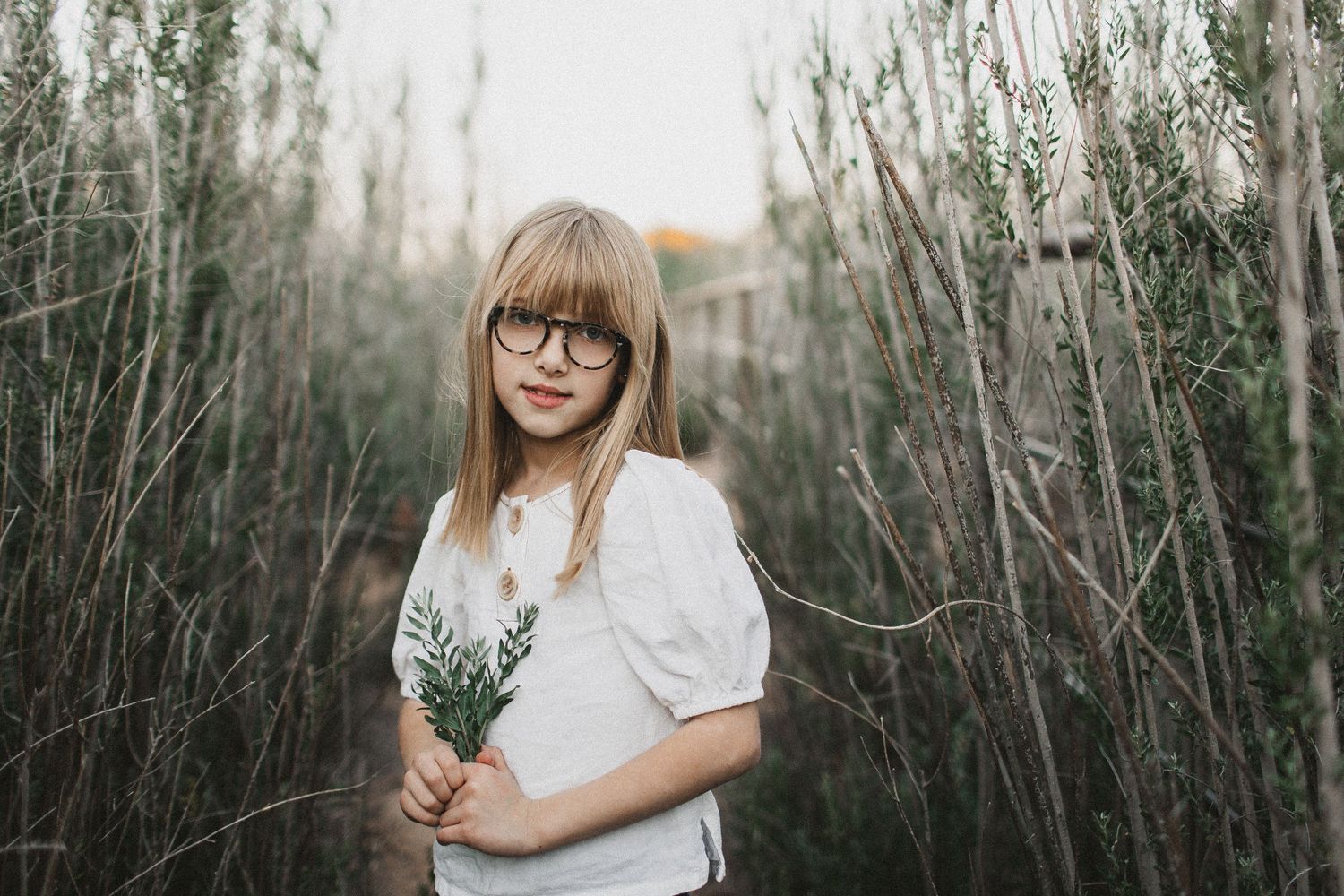 A young person in glasses and white top holds wildflowers while standing among tall grasses.