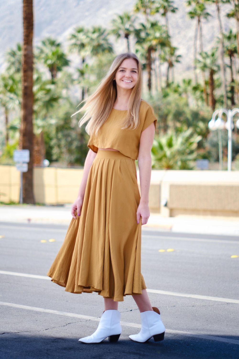 Flowing mustard yellow dress ensemble styled with white boots in desert palm setting.