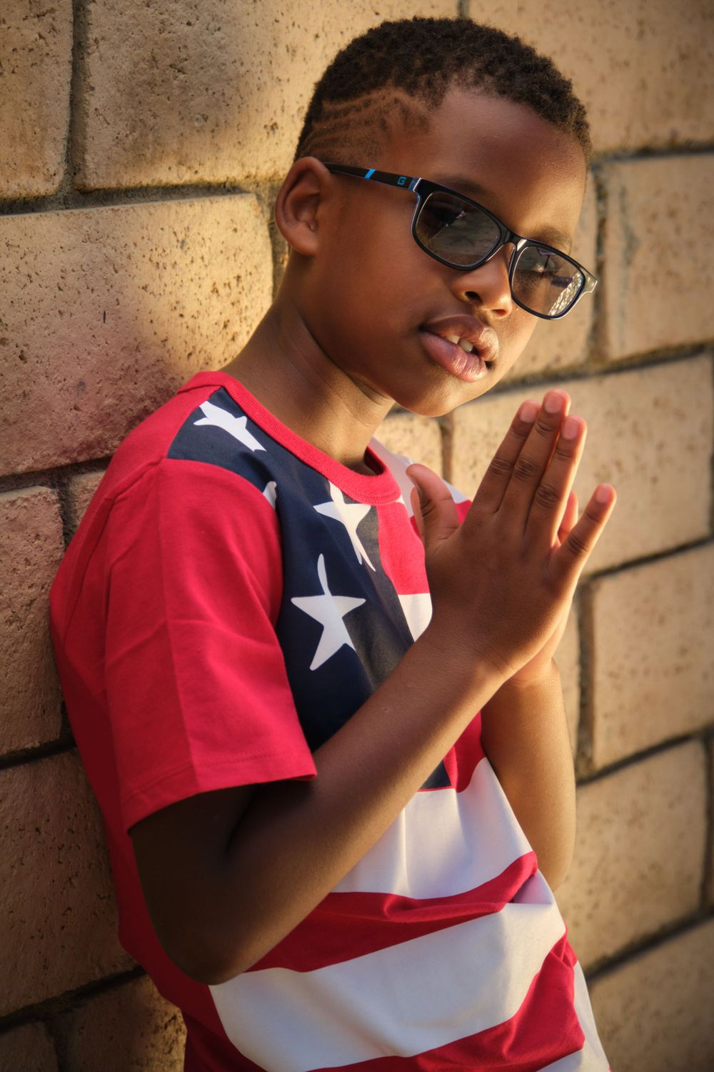 Youth in patriotic American flag t-shirt and glasses poses against brick wall.
