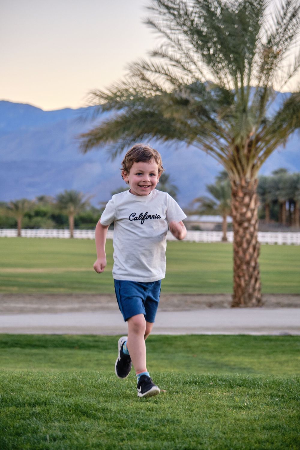 Child running on grass field with palm trees and mountains in background.