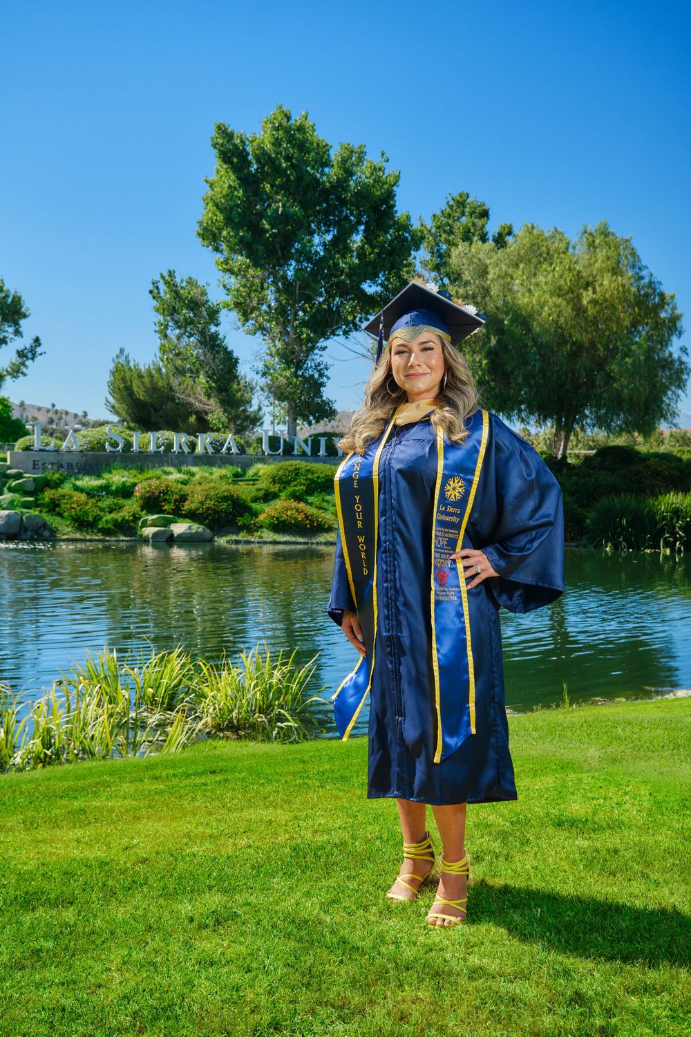 Graduate in blue cap and gown with gold sash poses by lakeside on sunny day.