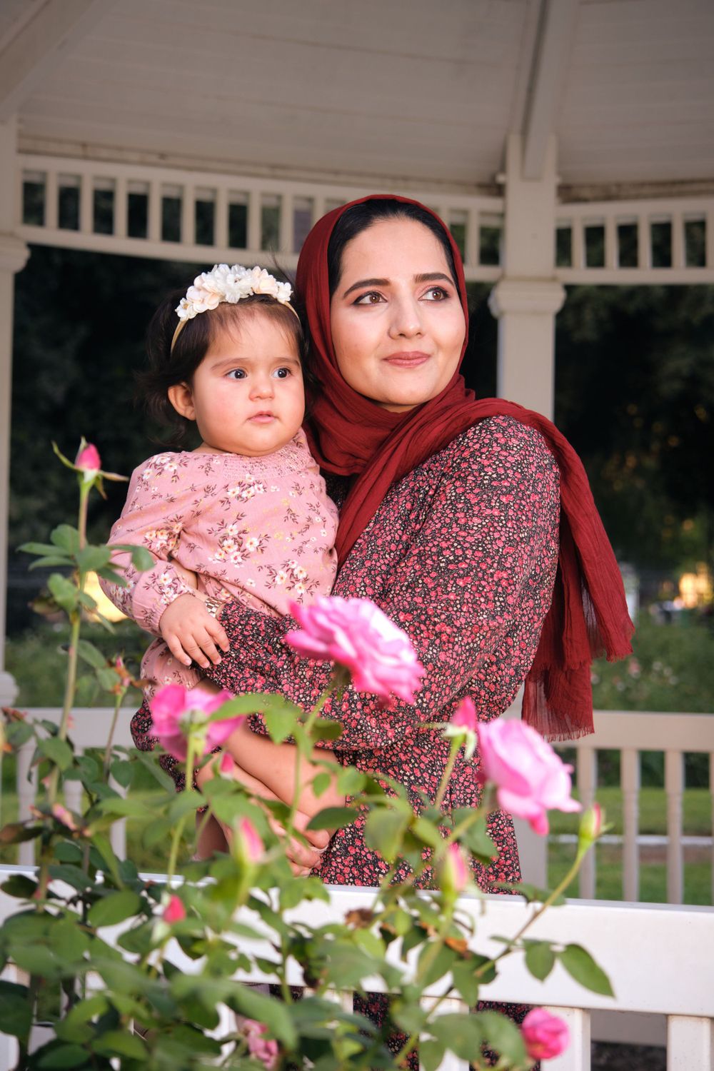Mother and child standing among blooming roses in white gazebo setting.