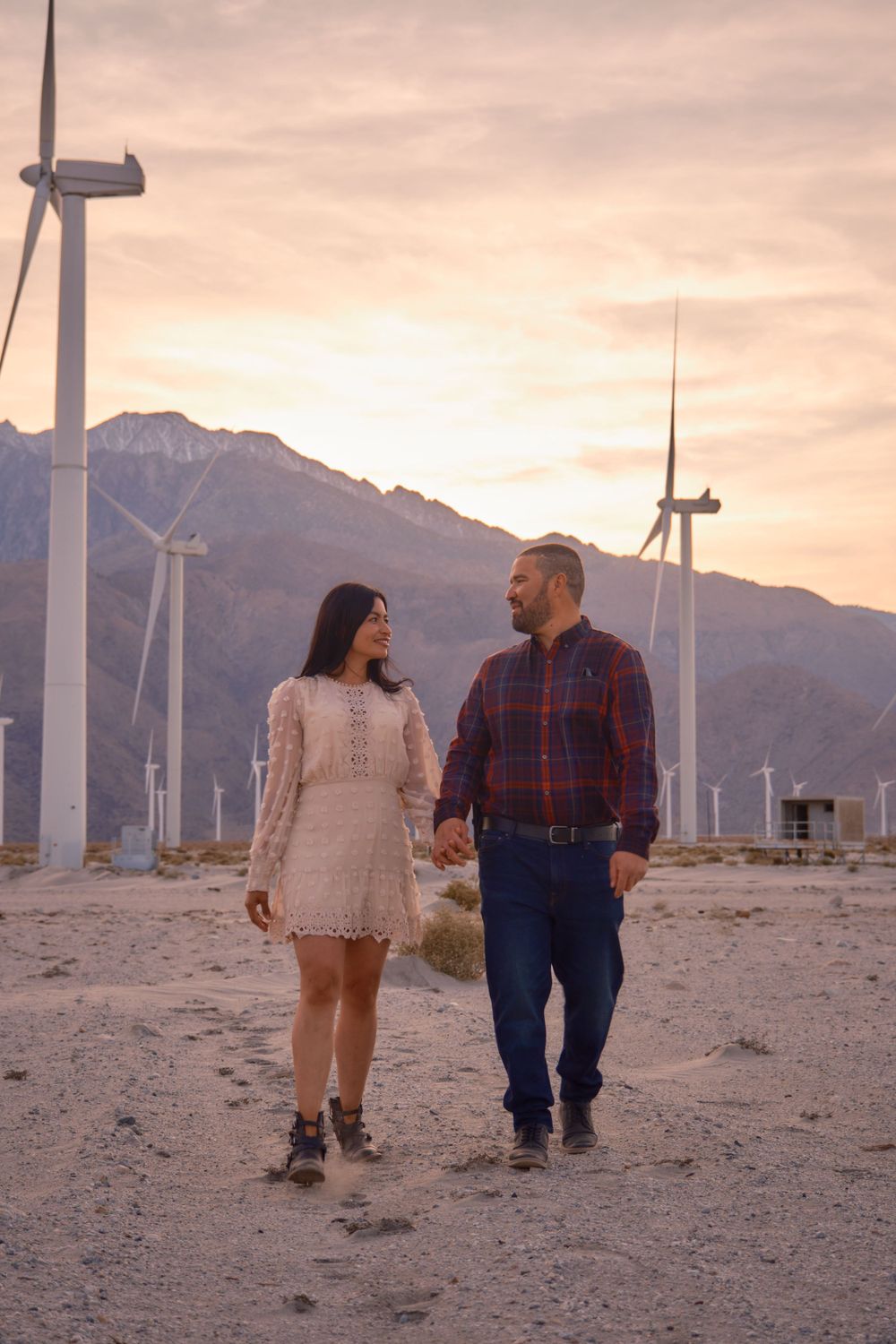 A couple walks hand in hand at sunset among wind turbines and mountains in the desert.