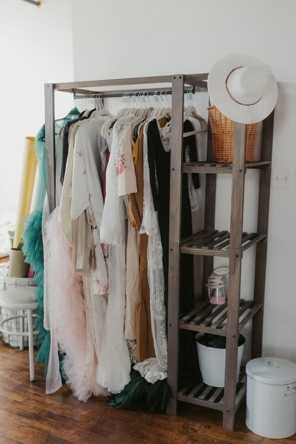 A wooden clothing rack displaying various light-colored dresses with a white hat and storage bins in a bright room.