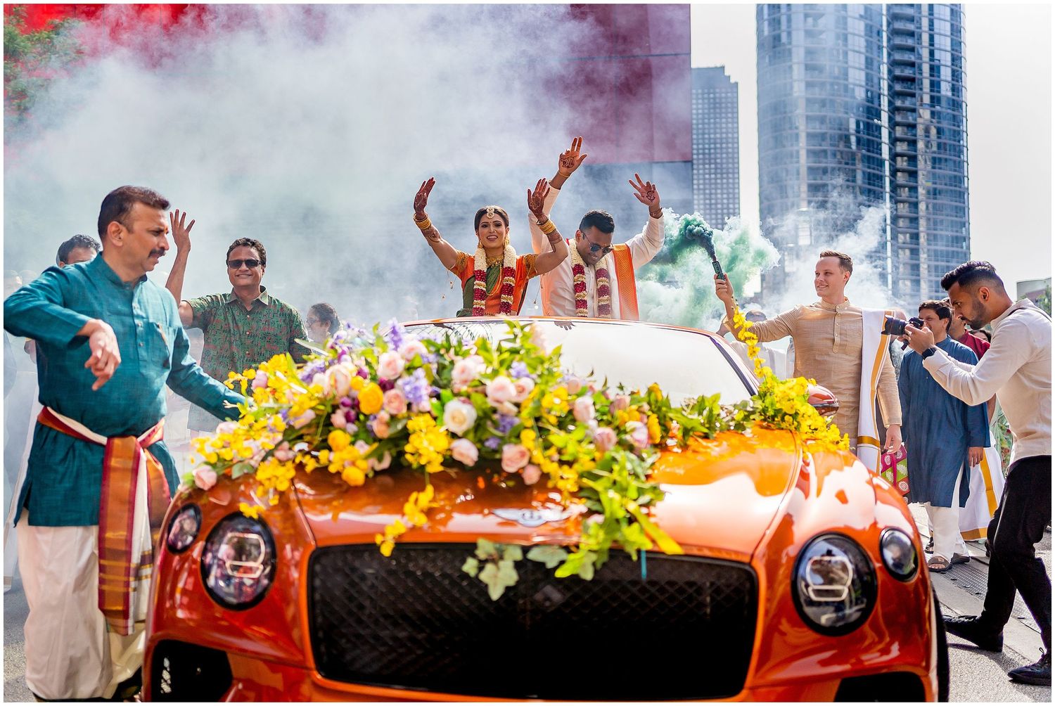 Orange Bentley decorated with yellow flowers amidst celebratory smoke in an urban setting.