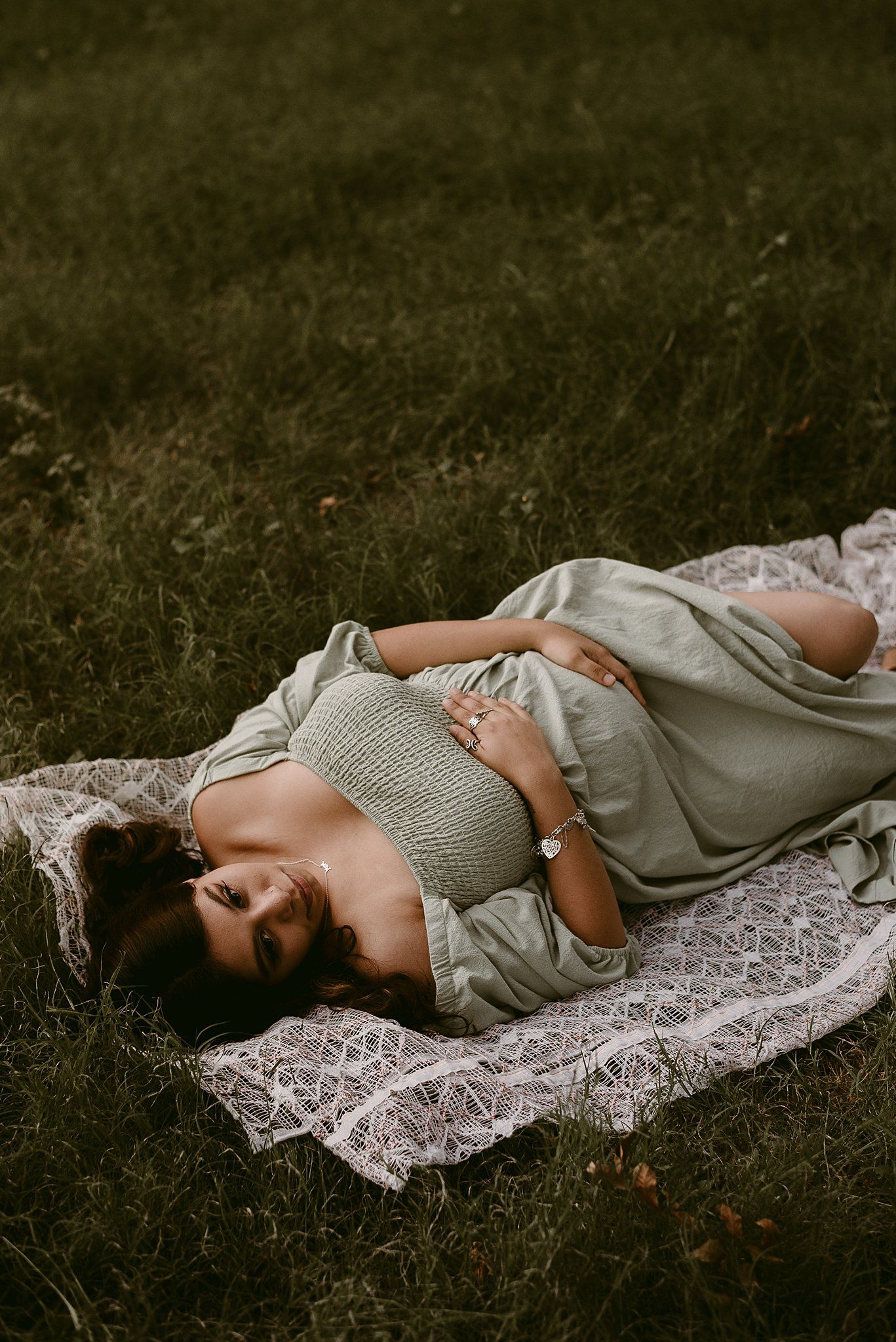 Maternity portrait of woman in grey dress lying on lace blanket in garden setting.