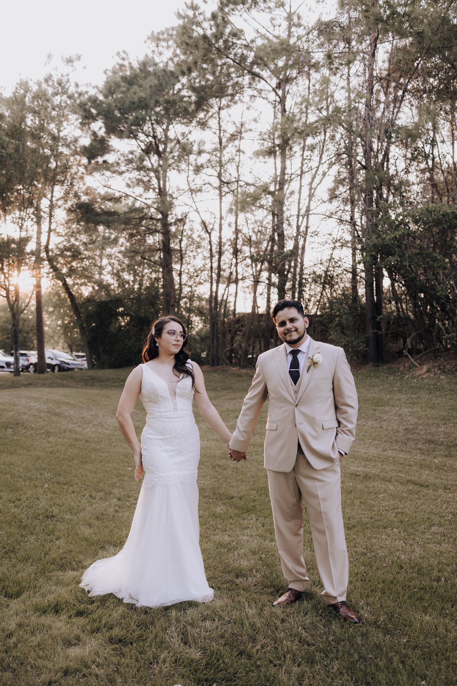 Bride in white lace gown and groom in beige suit hold hands during outdoor forest wedding at sunset.