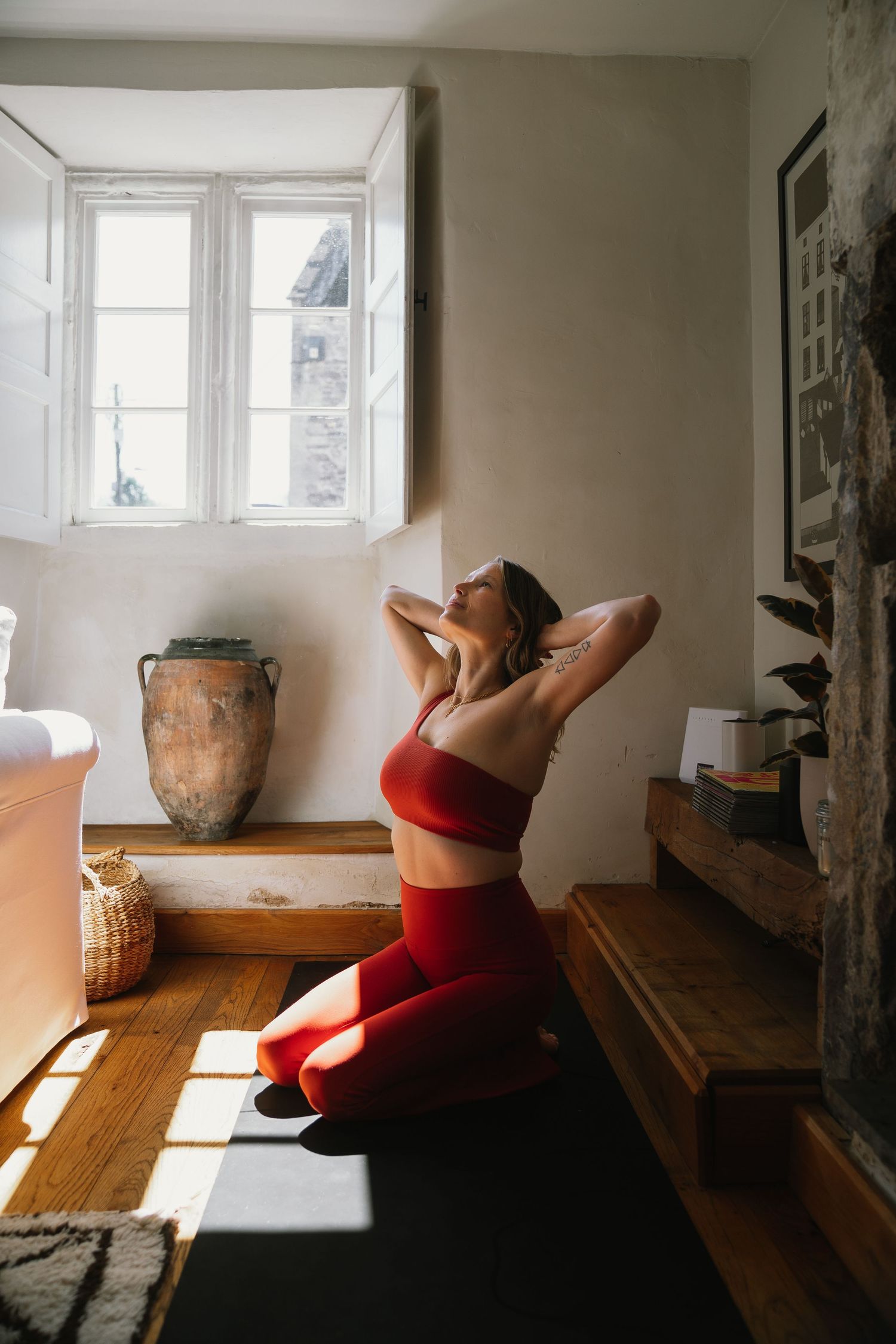 A person in red workout attire stretches in a sunlit room with wooden floors and a decorative clay pot.