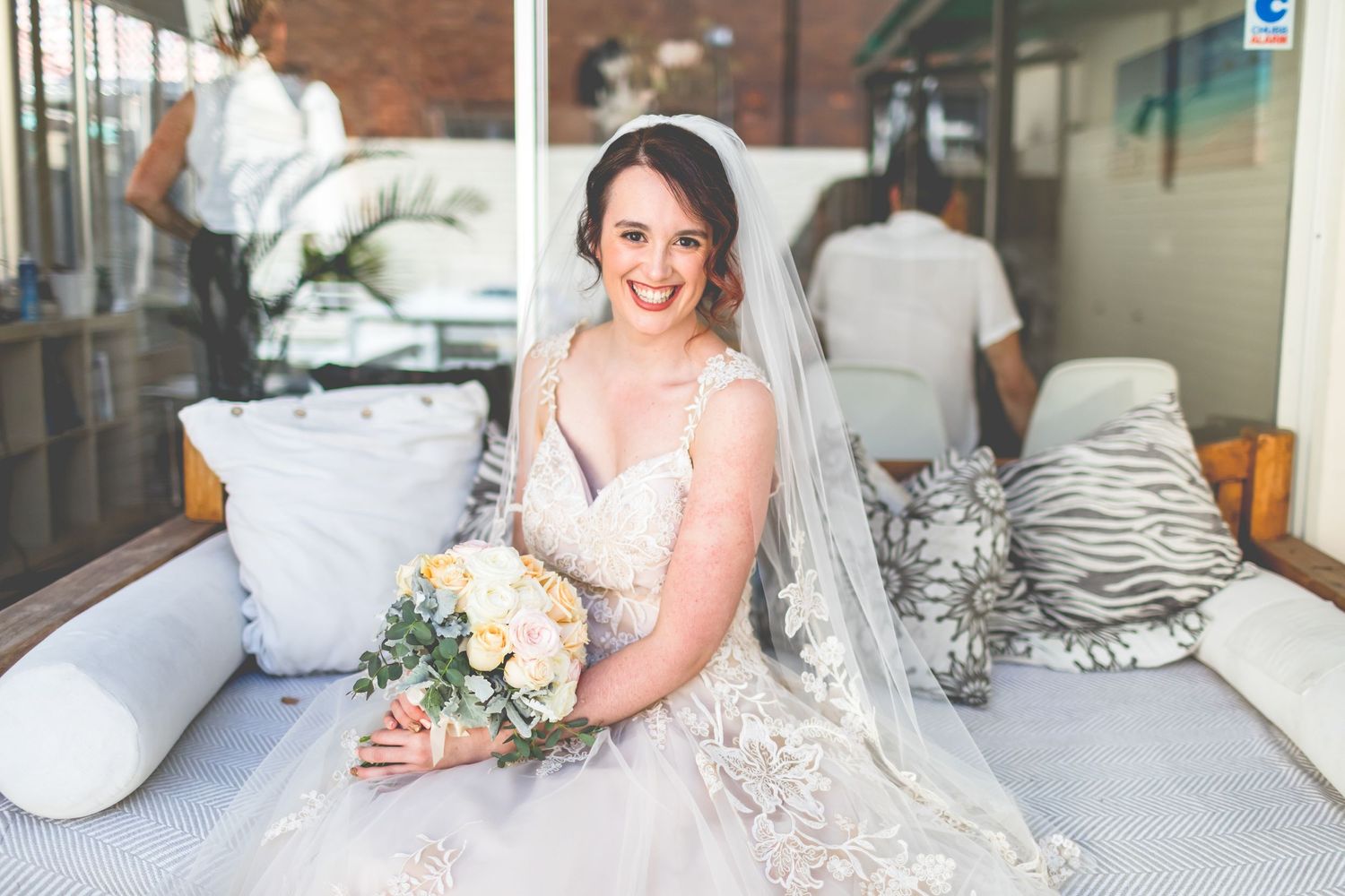 Bride in lace wedding dress holding bouquet while sitting on white couch.