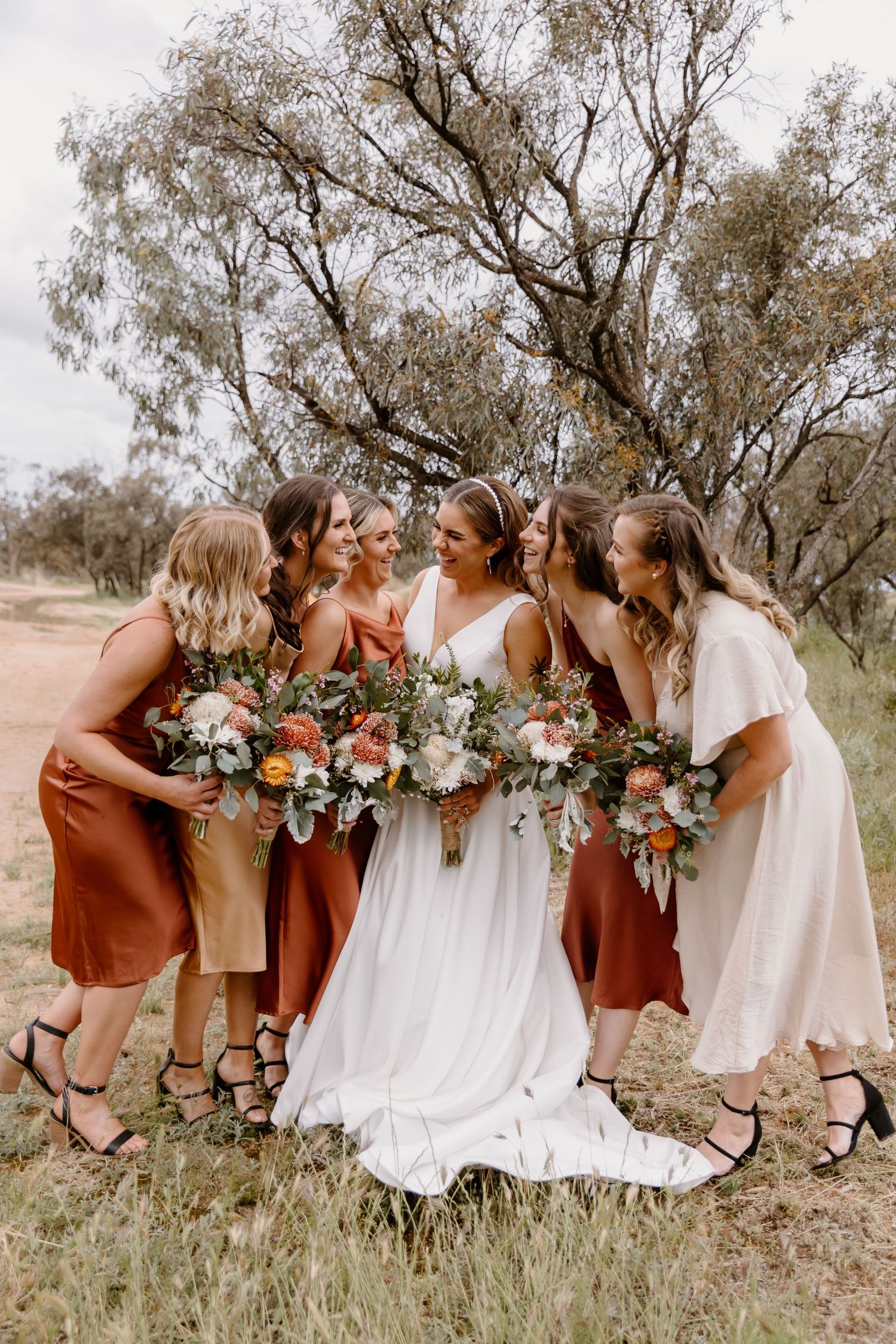 A bridal party in terracotta and white dresses huddles together with bouquets in an outdoor setting.