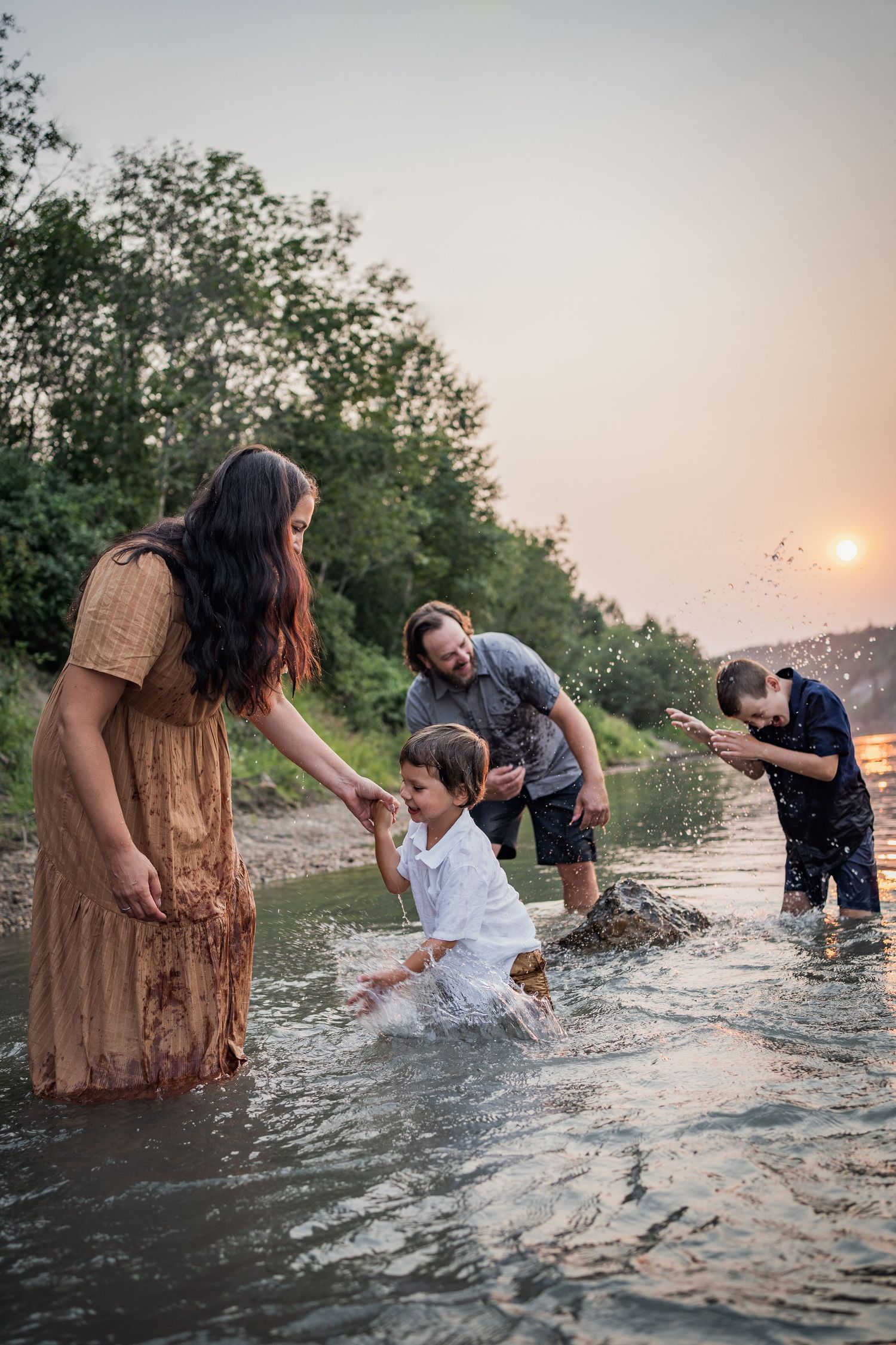 Family playing and splashing in shallow water at sunset near a wooded shore.