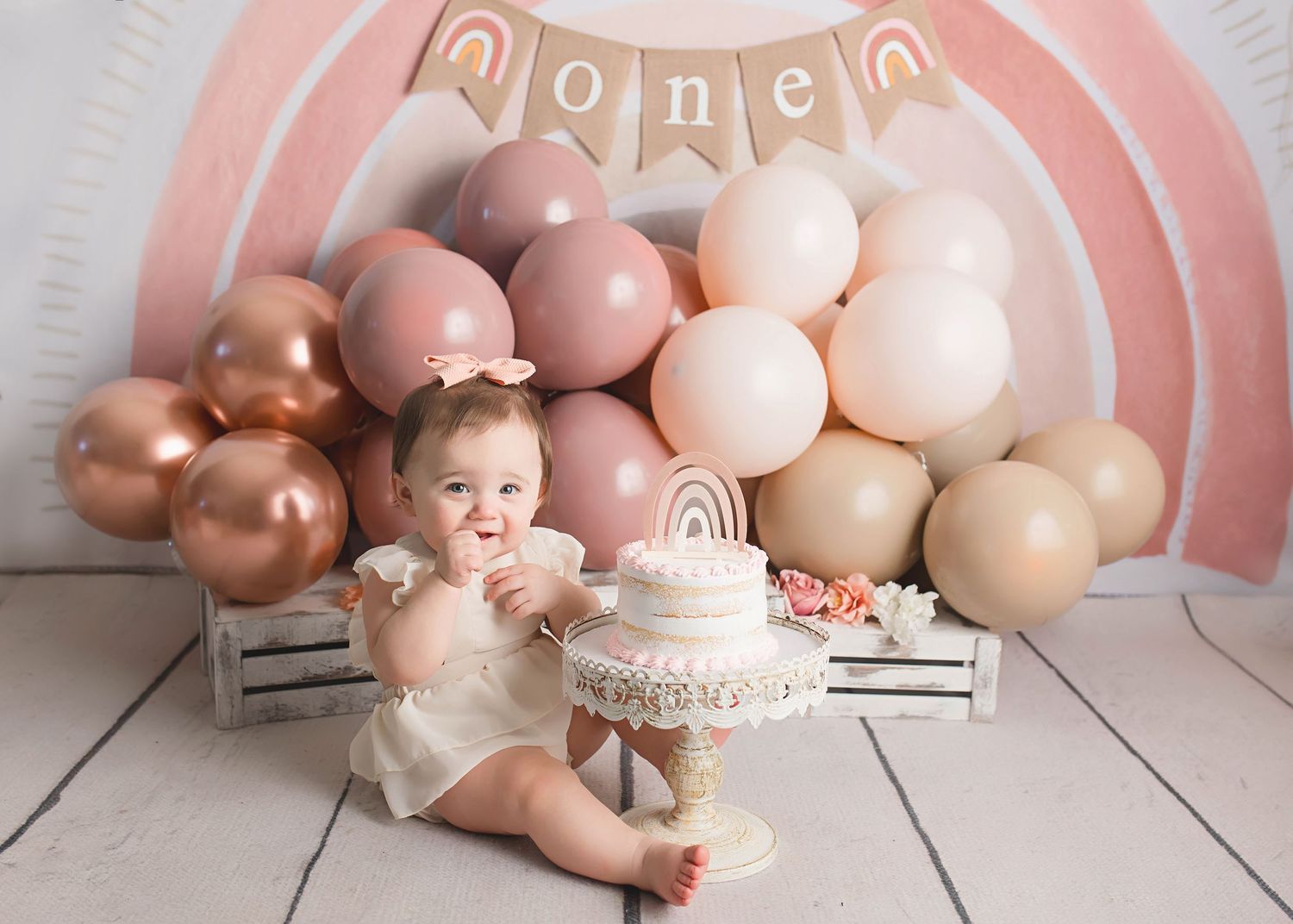 Baby milestone photo featuring pink and rose gold balloon backdrop with celebration cake.