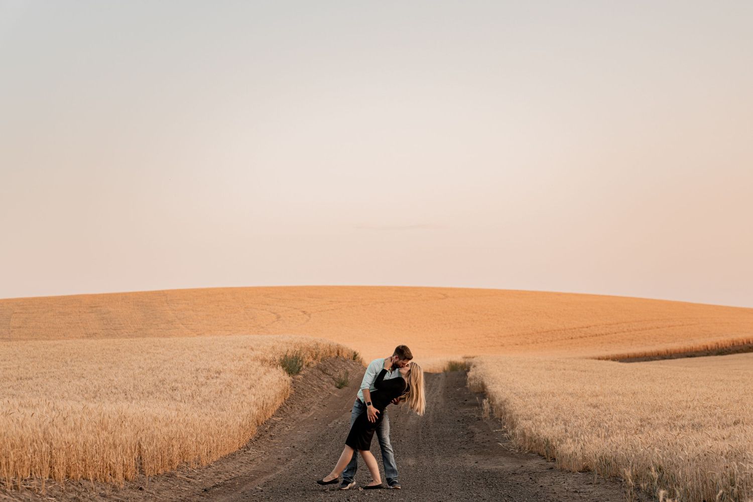 Two people embracing on a dirt path through golden wheat fields at sunset.