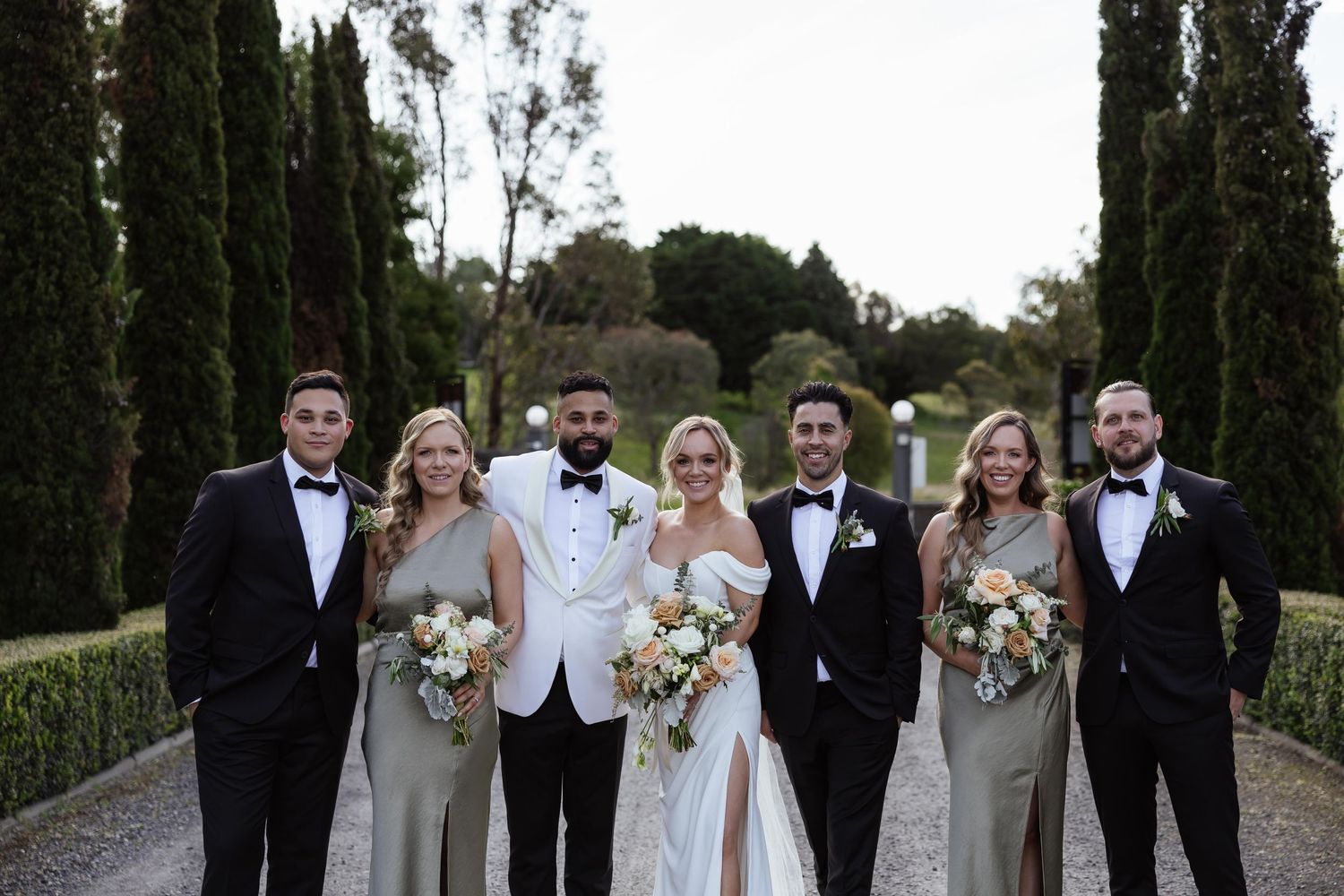 Wedding party poses for photos on a tree-lined path during an outdoor celebration.