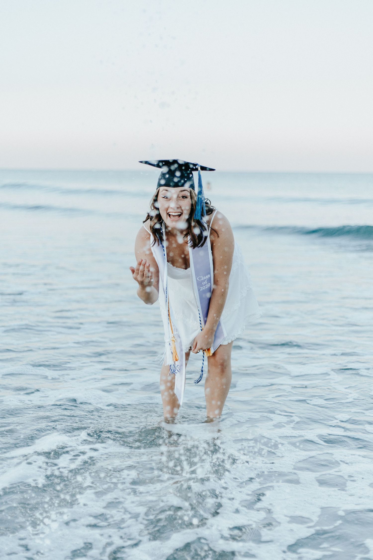 Graduate wearing cap splashing in ocean waves wearing white dress.