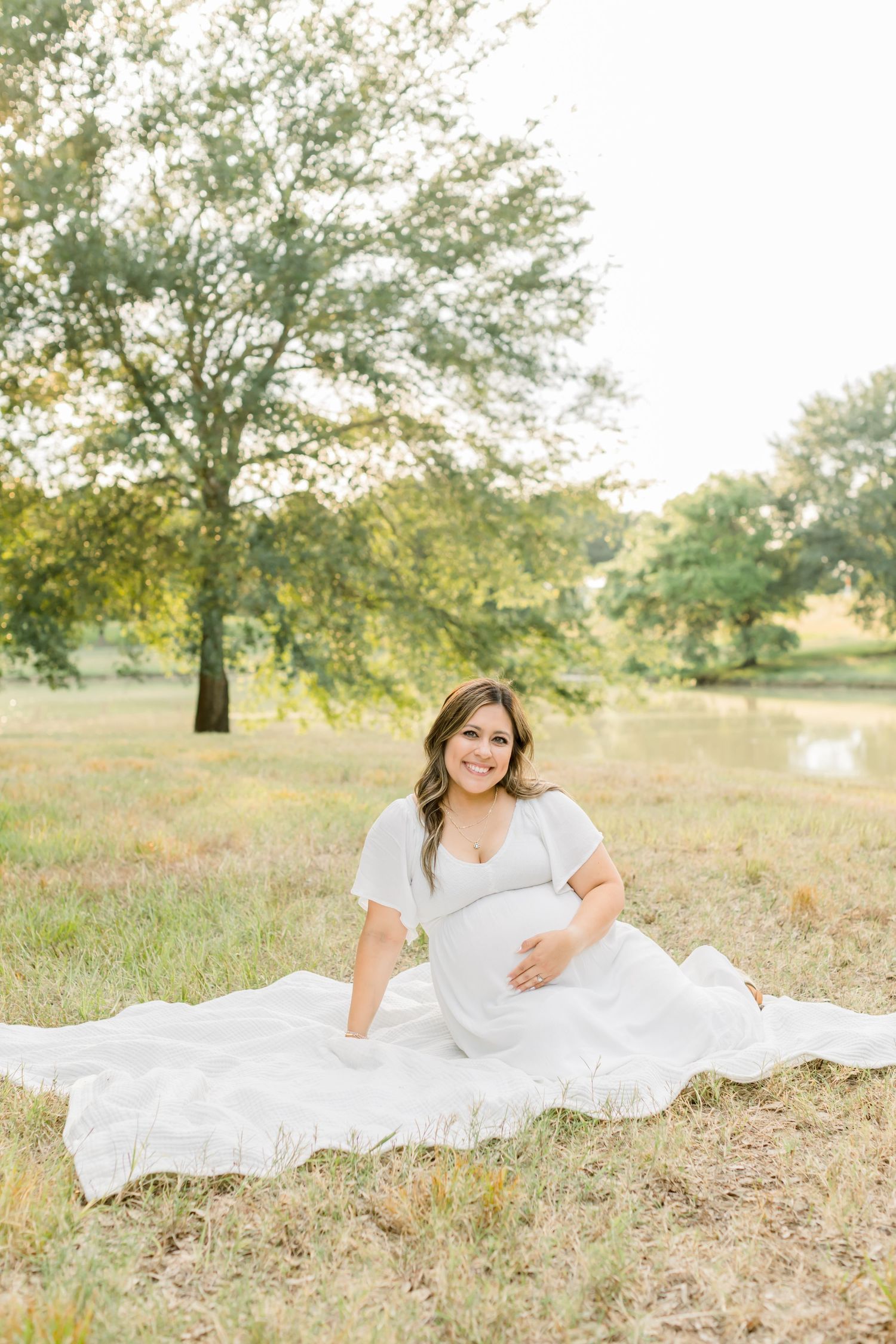 Expectant mother in white dress sits gracefully on a white blanket in a sunny field.