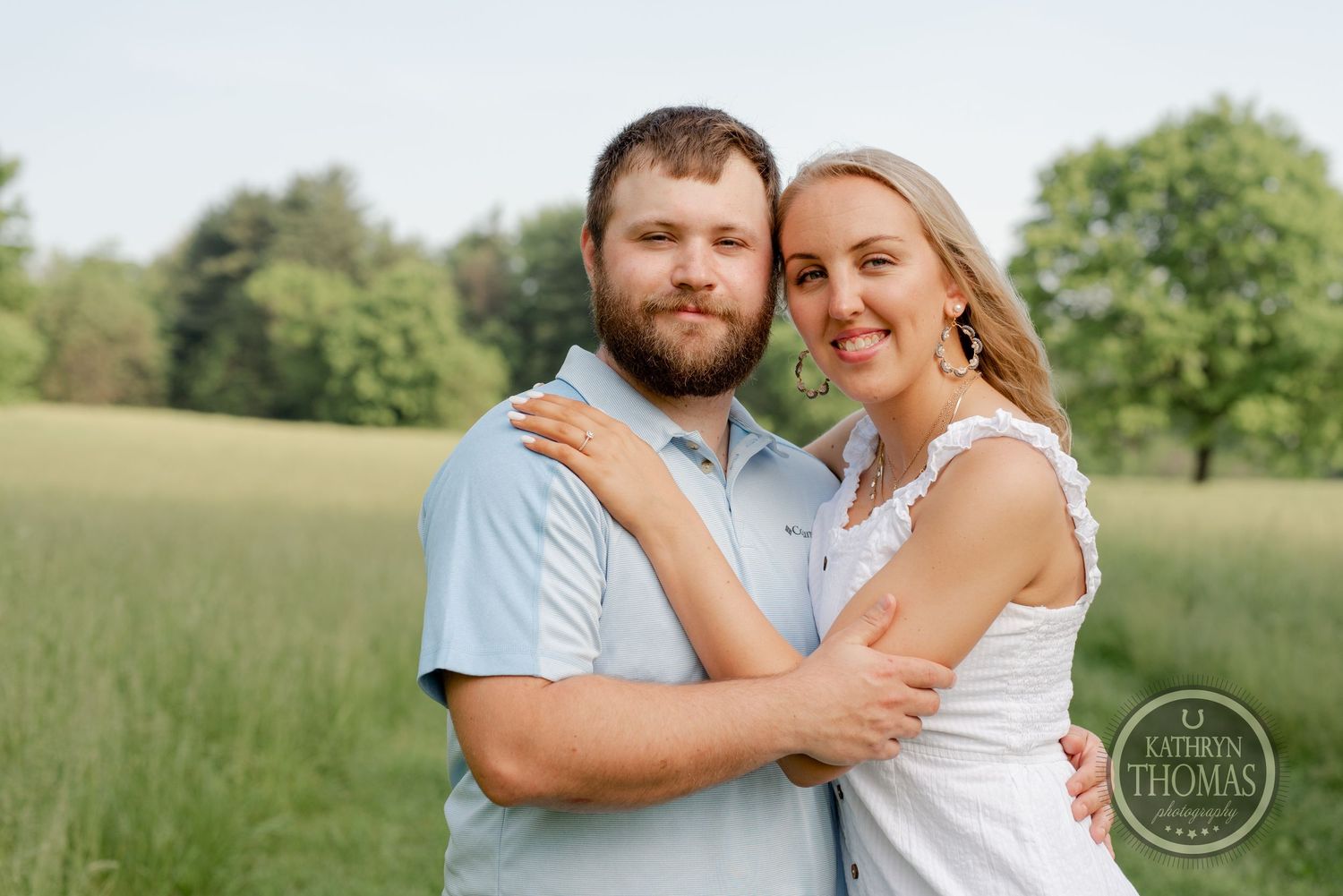 A couple embraces in a sunny meadow wearing light colored clothing during a summer photoshoot.