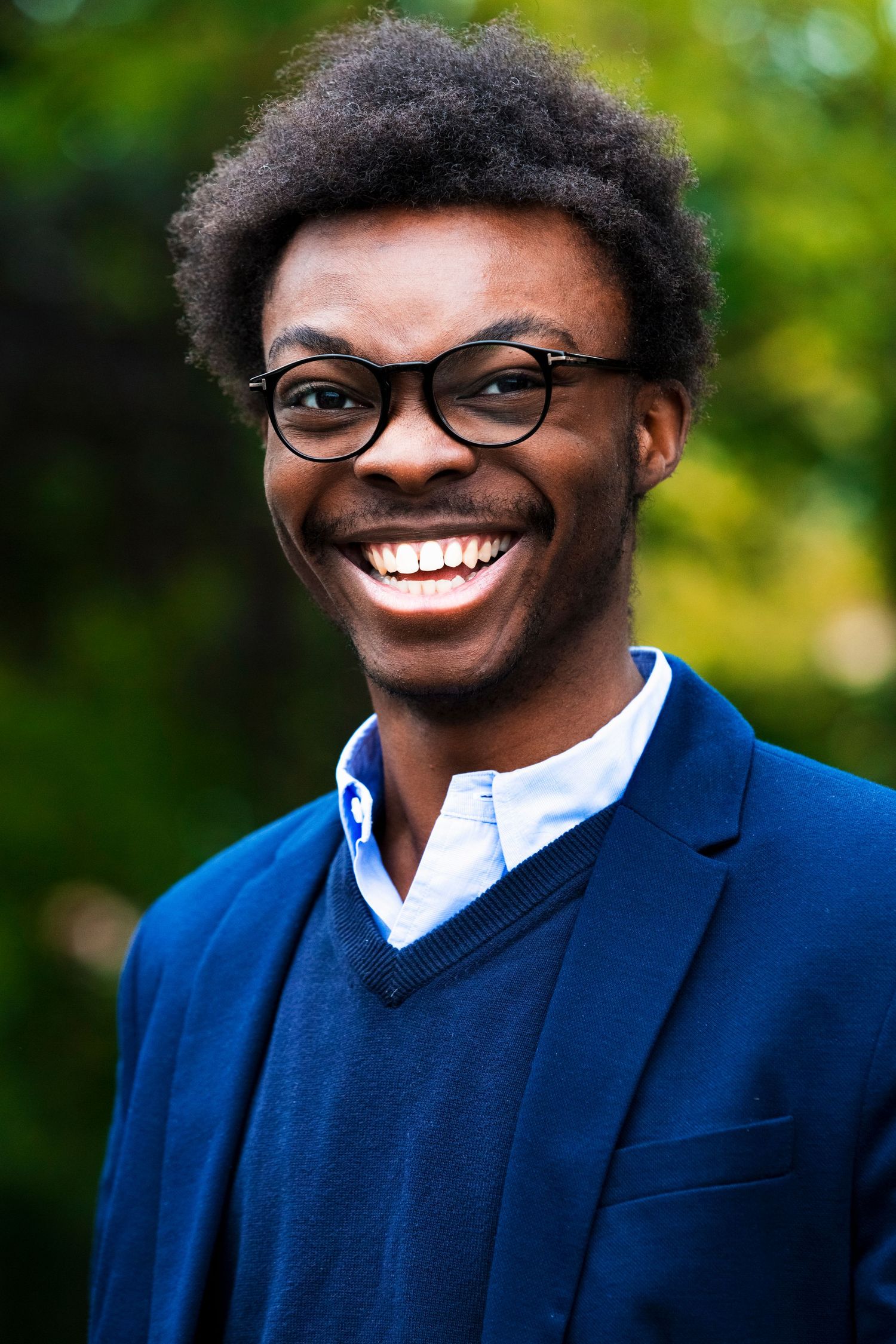 Professional headshot of person in navy sweater and glasses with natural background.