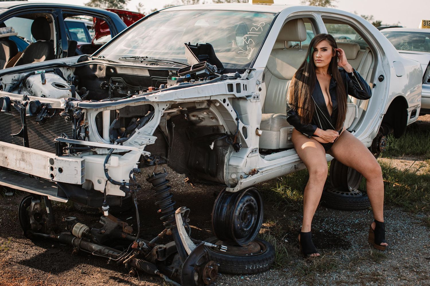 Woman in black jacket posing next to dismantled car in junkyard.