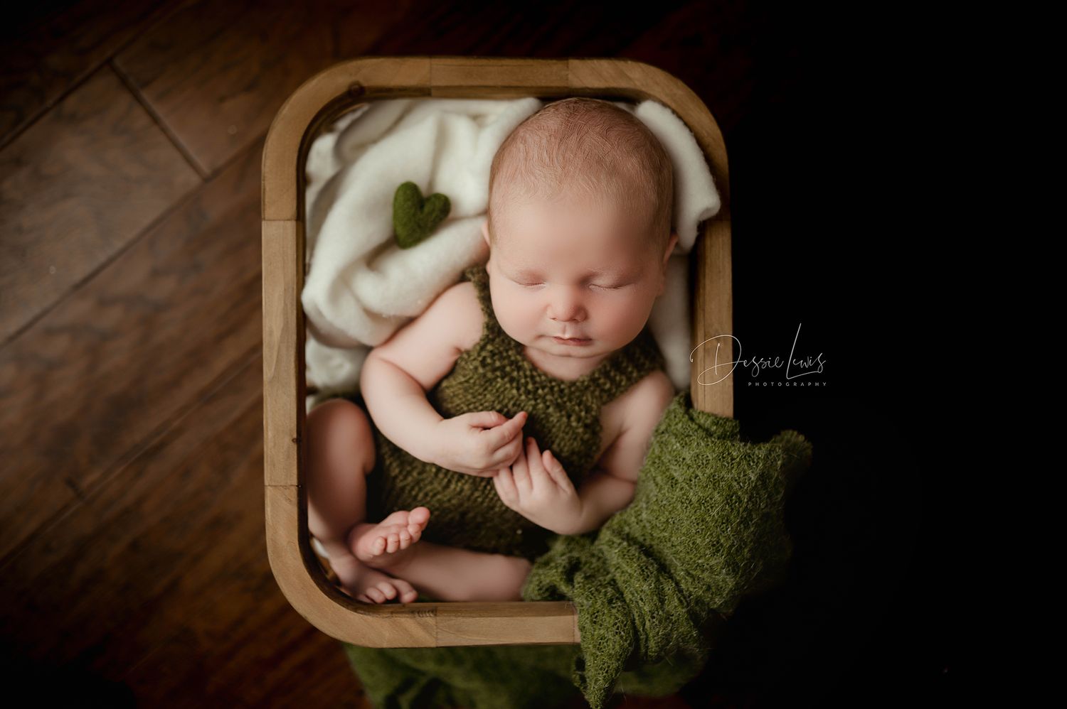 Sleeping newborn in wooden basket with green blanket on hardwood floor.