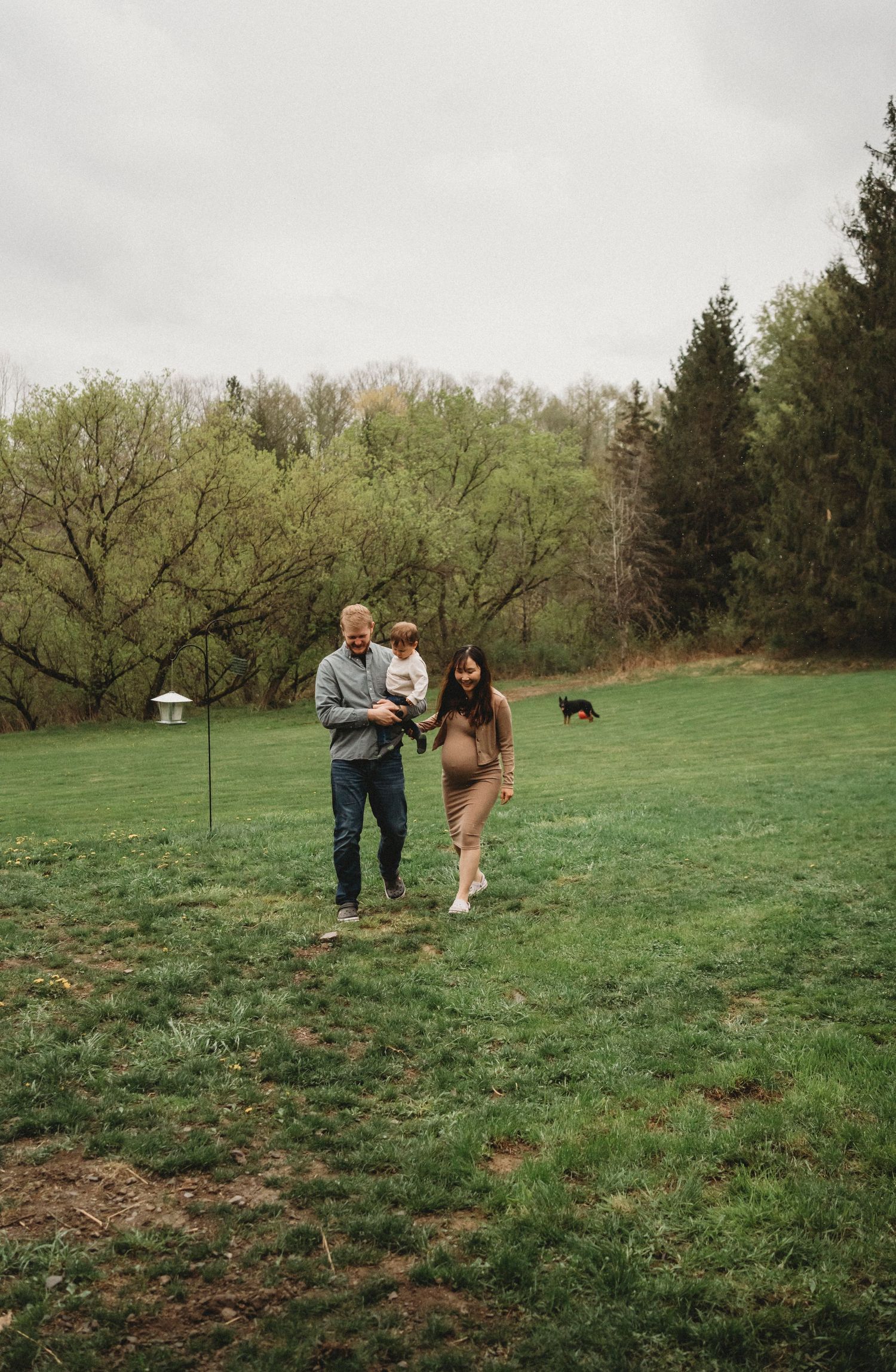 A couple enjoys a playful moment on a grassy field with trees in the background and a dog visible in the distance.