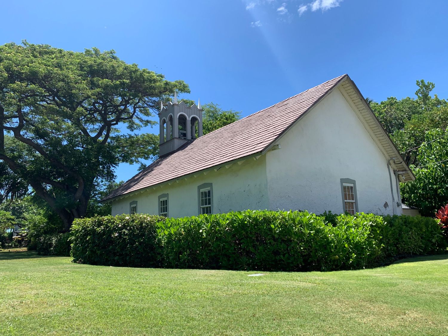 A small white Hawaiian church with a red tile roof surrounded by lush green trees and lawn.