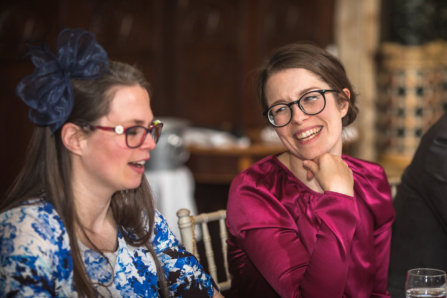 Two wedding guests in colorful attire share a laugh at reception table.