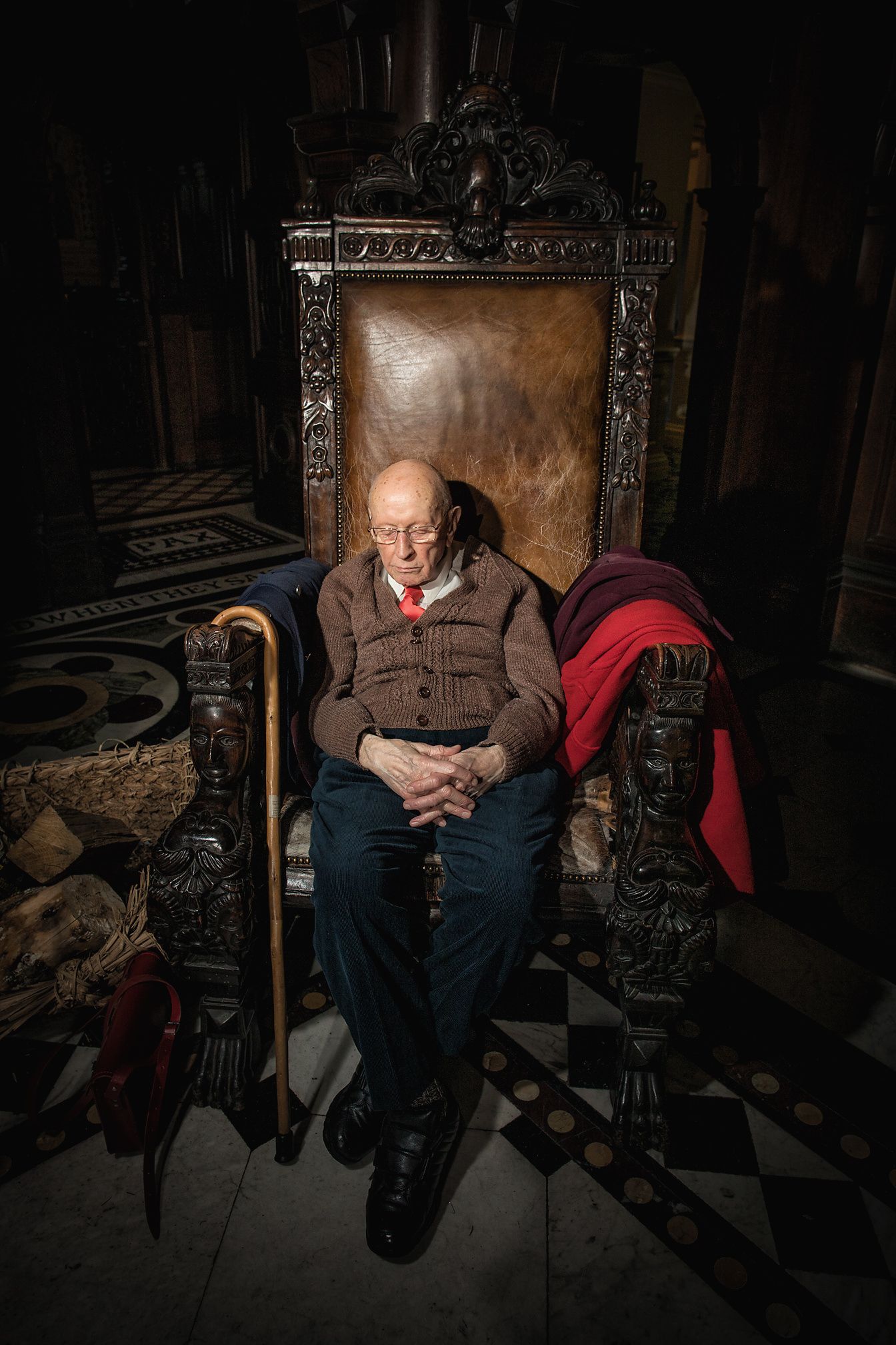 Elderly person rests in ornate carved wooden throne chair wearing cardigan sweater.