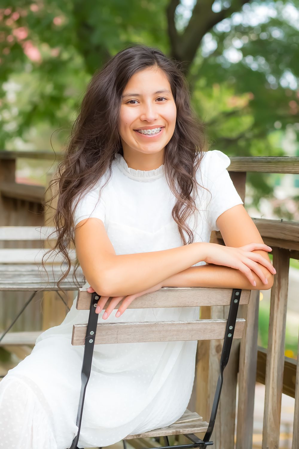 A person in a white dress sits casually on an outdoor patio chair with lush greenery in the background.