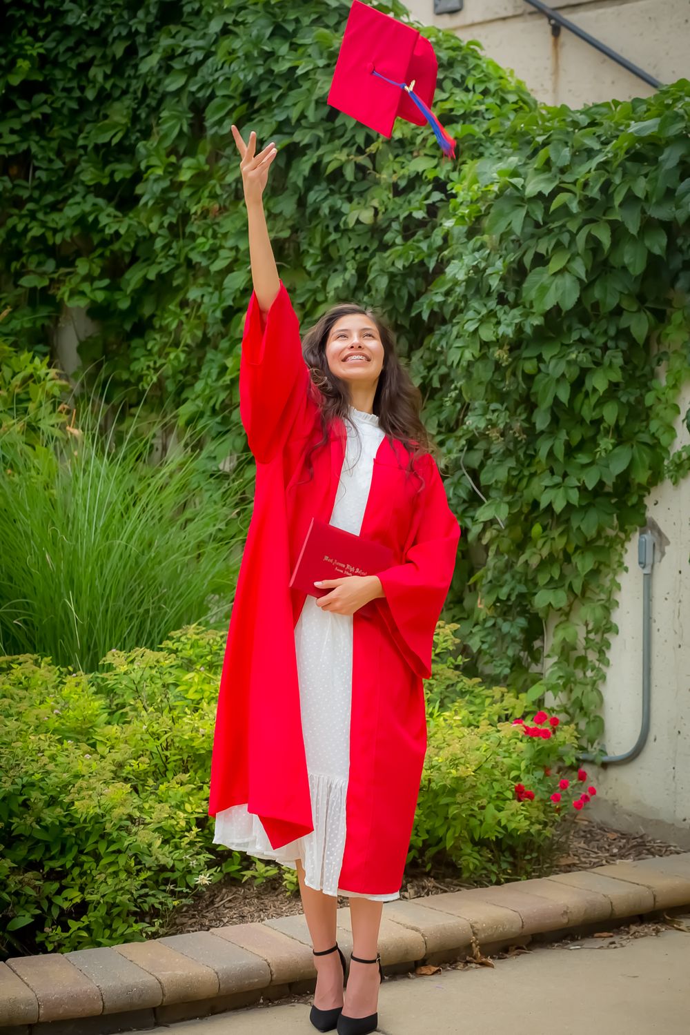 A graduate in red academic regalia throws their cap in celebration against an ivy-covered wall.