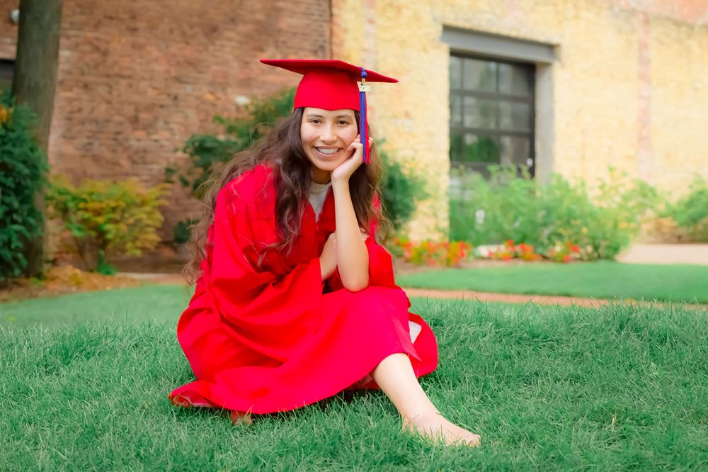 A graduate in bright red cap and gown sits on grass during a celebratory photoshoot.