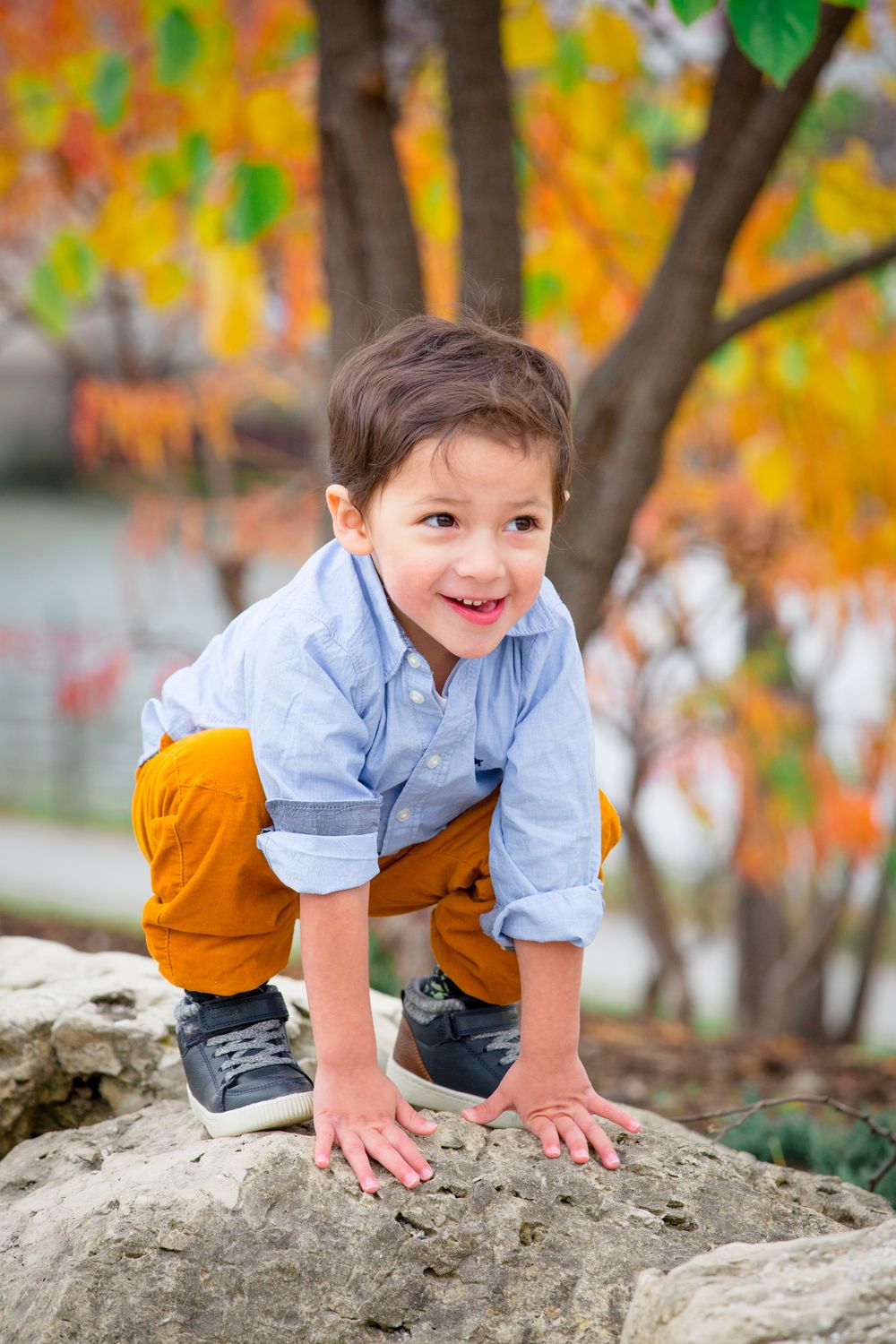 A playful child in yellow pants and blue shirt climbs on a large rock during fall season.