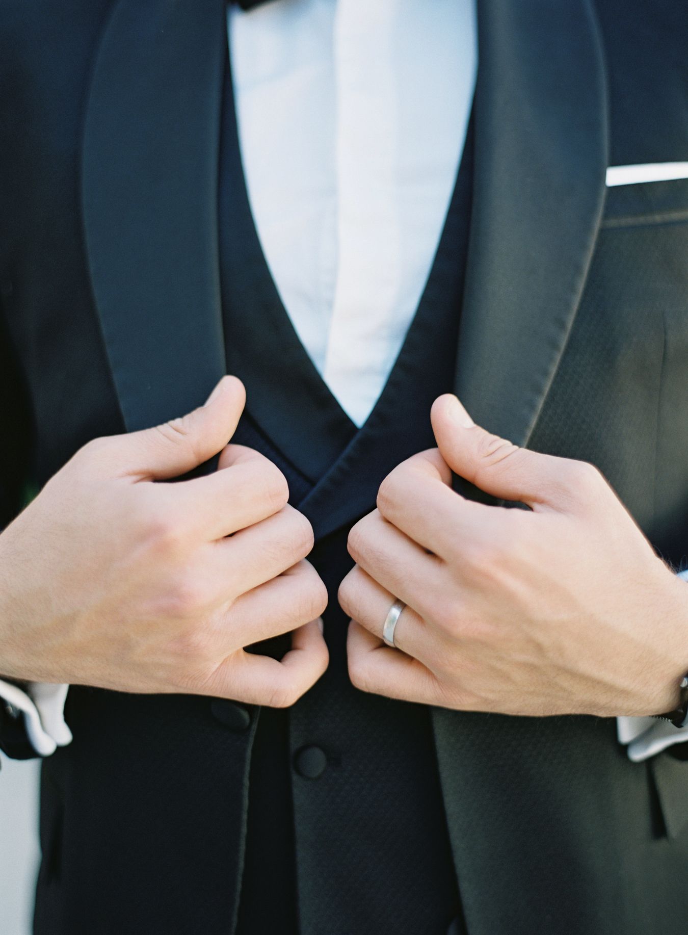 Close-up of hands adjusting elegant black tuxedo lapels with wedding ring detail.