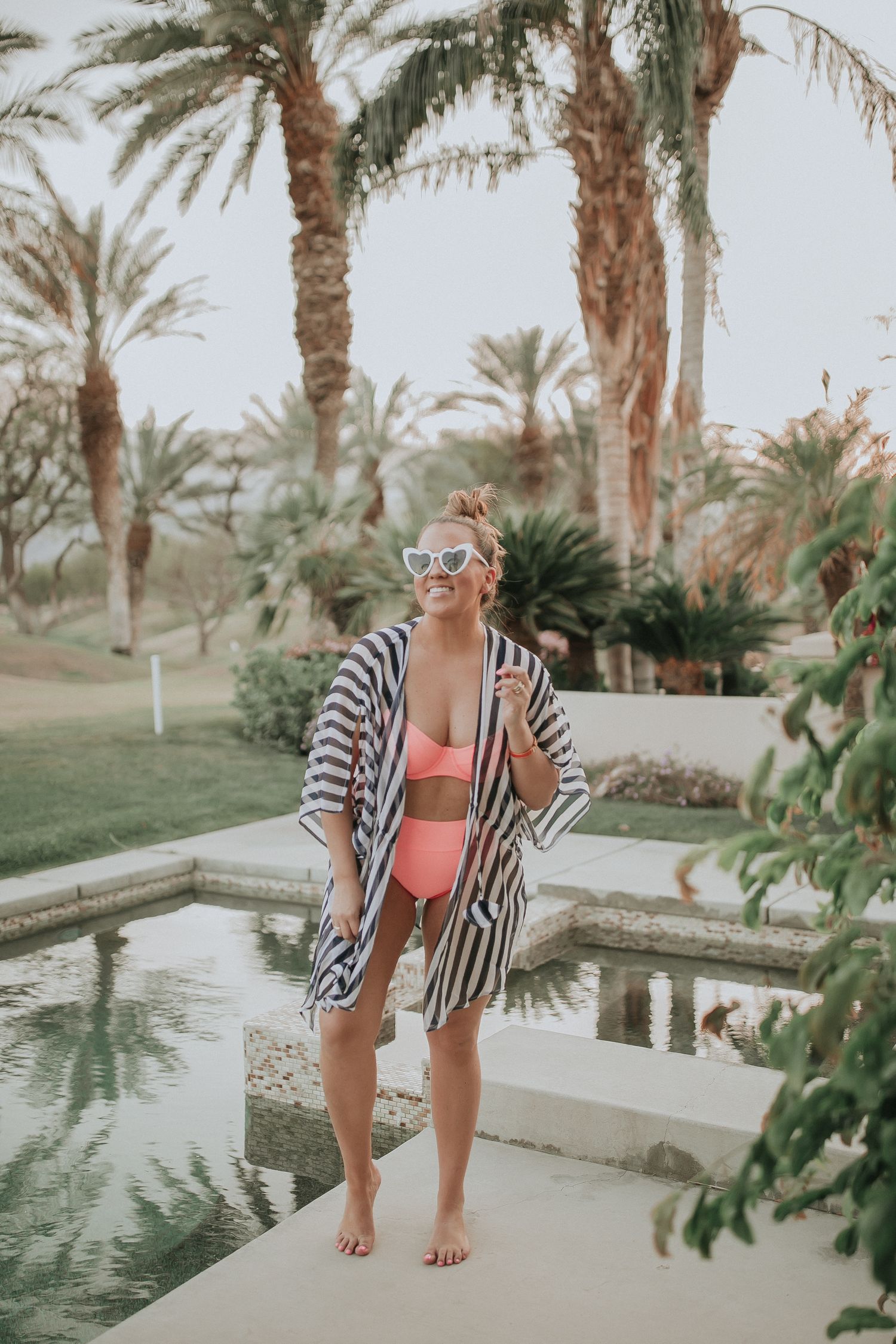 Person in striped cover-up and swimwear poses by palm-lined pool in tropical resort setting.