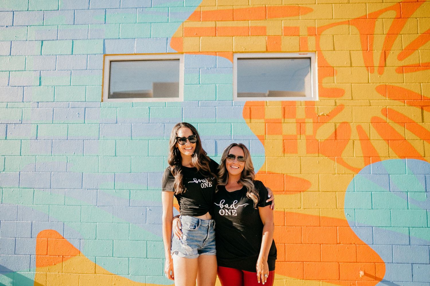 Two people pose against colorful geometric mural wall with orange and mint green patterns.