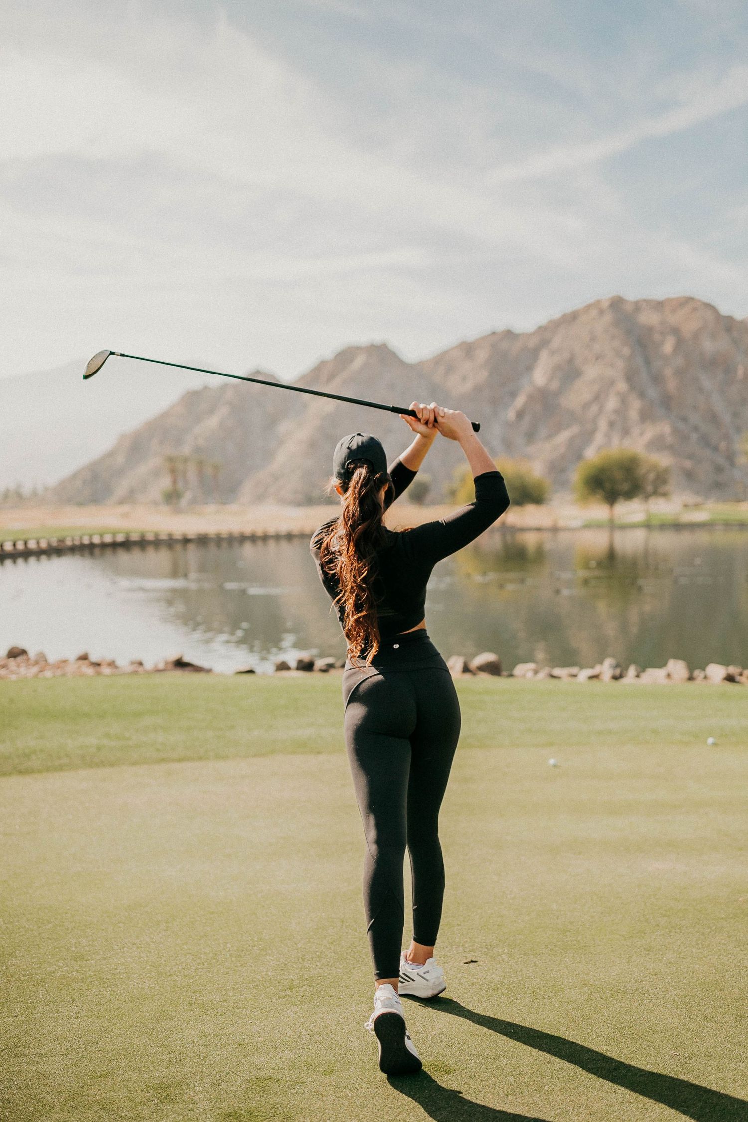 Golfer in dark athletic wear takes a swing on course with mountains and lake in background.
