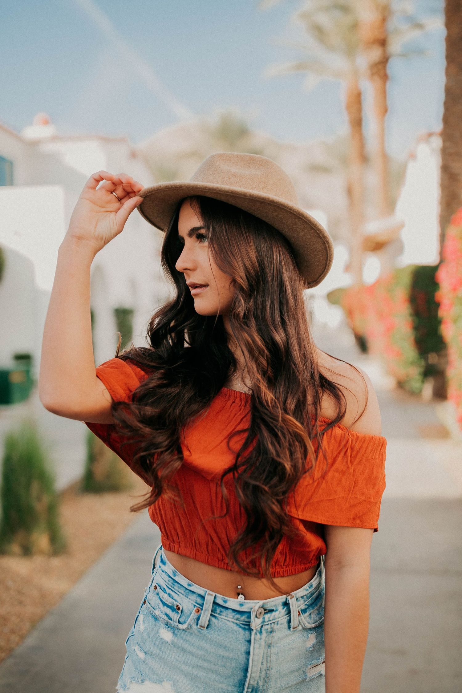 Fashionable woman in red crop top and denim wearing a beige hat poses on a sunny street with palm trees.
