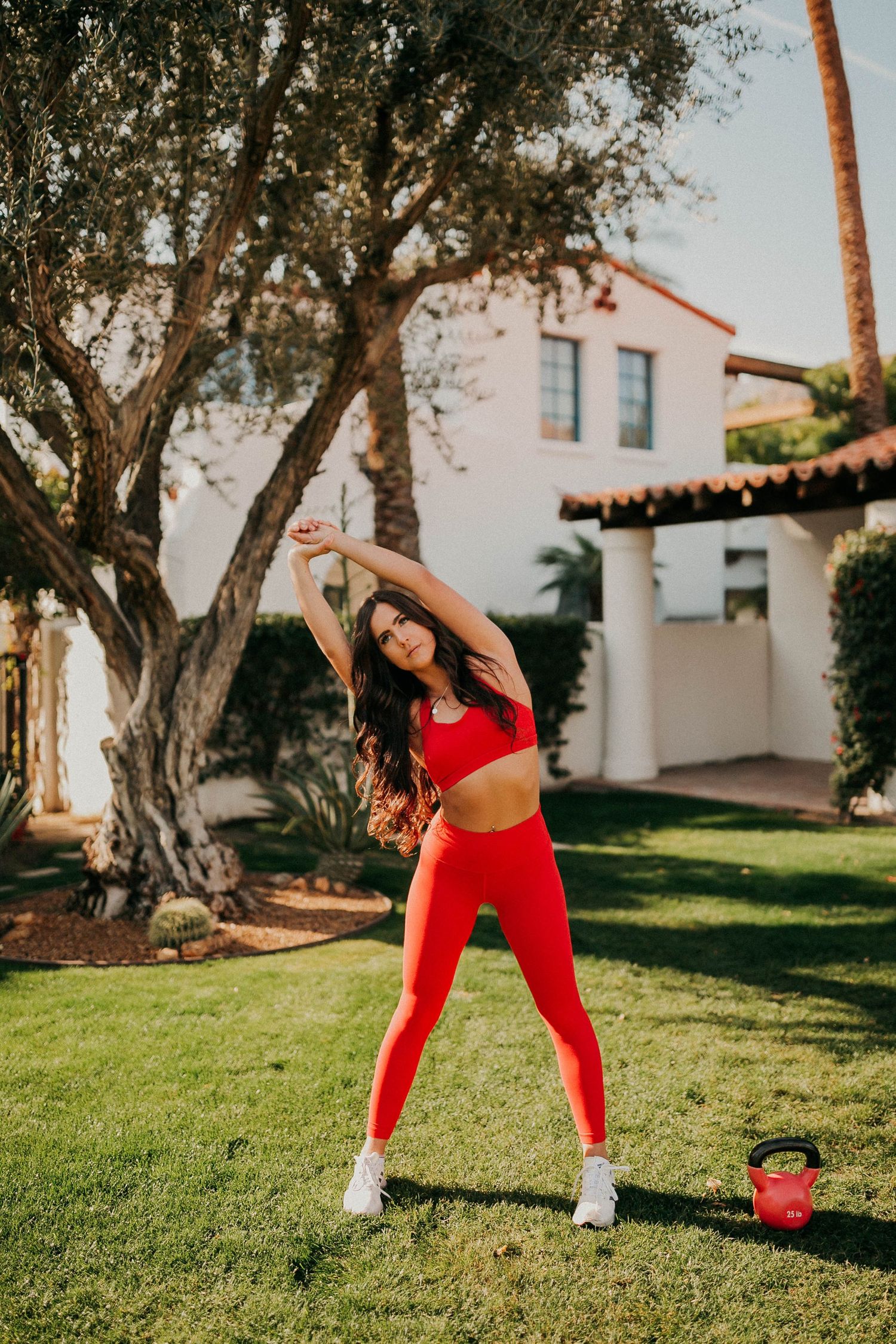 Woman in red athletic wear stretches outdoors on lawn with Spanish-style building in background.