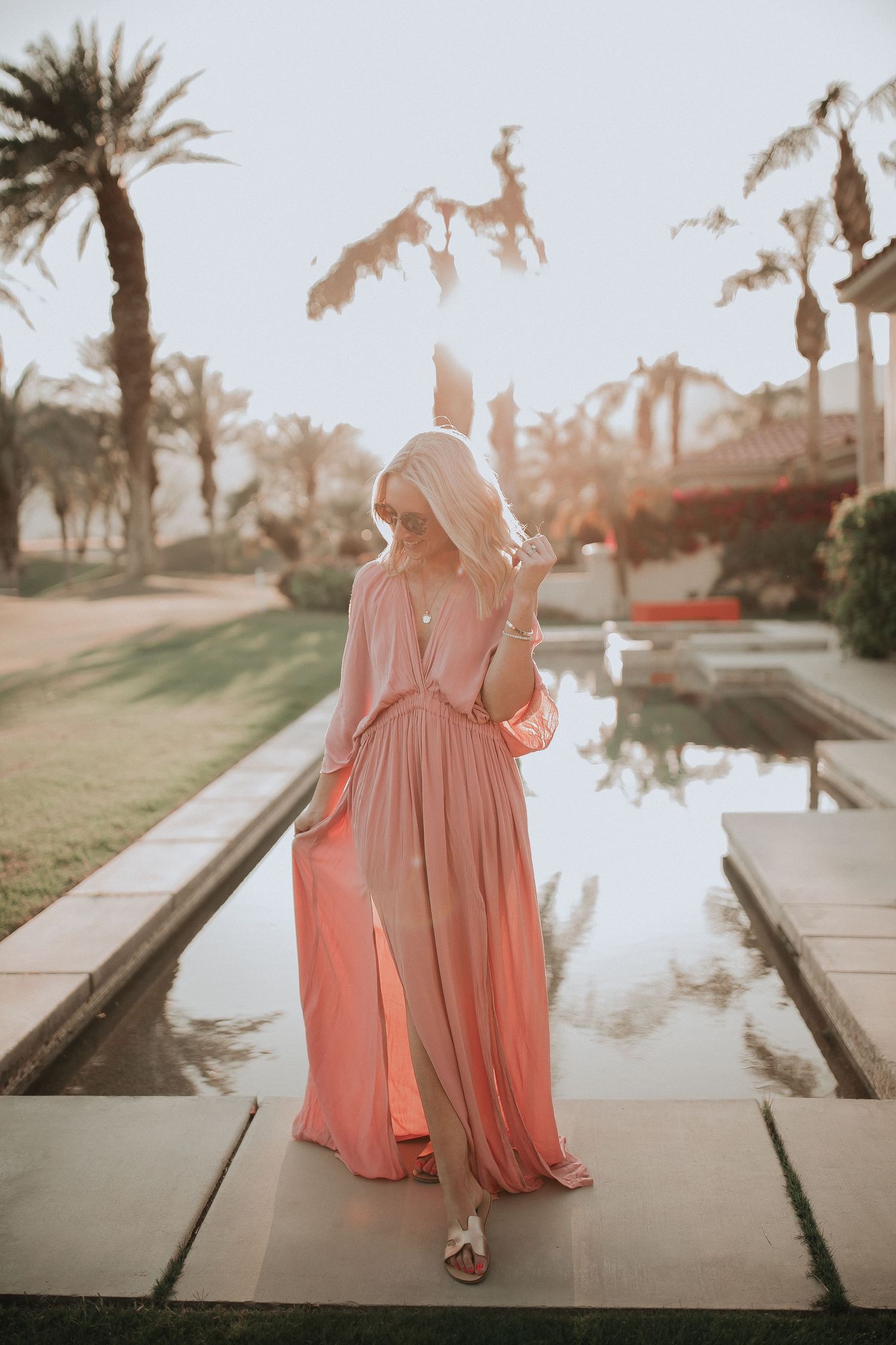 Woman in flowing pink dress stands by reflecting pool with palm trees at sunset.