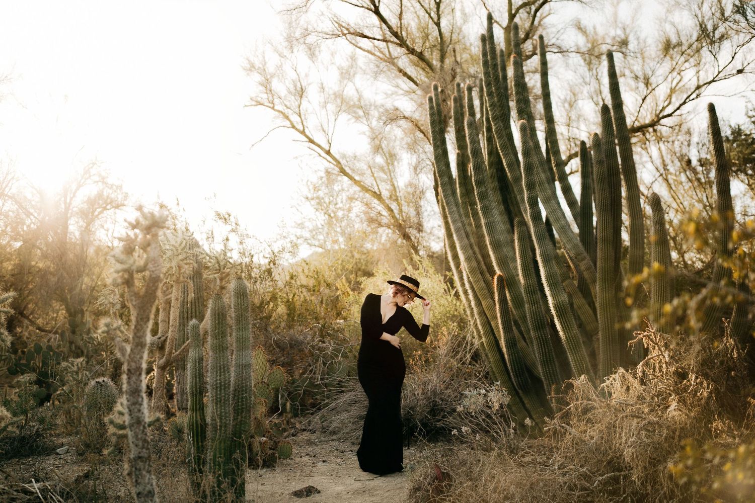 Silhouetted figure in long dress poses among desert cacti at golden hour.