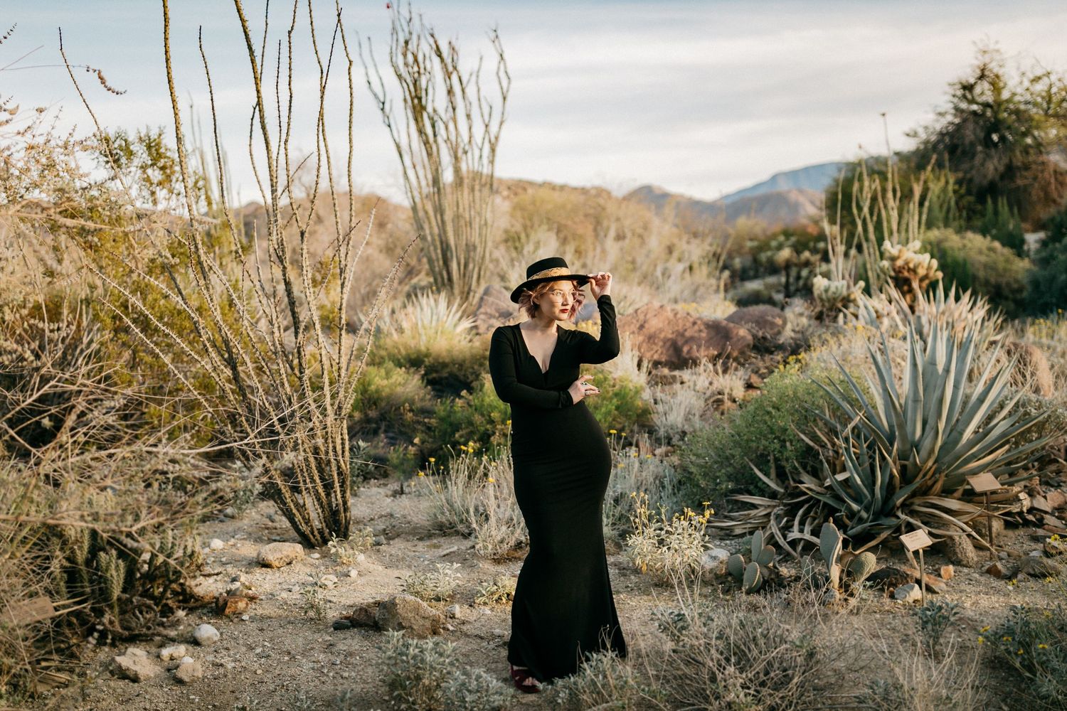 Person in black maxi dress and hat poses in desert landscape with cacti.